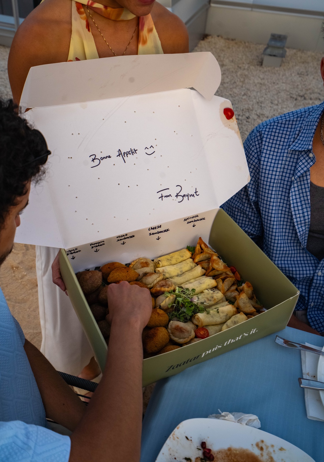 Guests opening a BÁYRŪT catering box with a handwritten Bon Appétit note