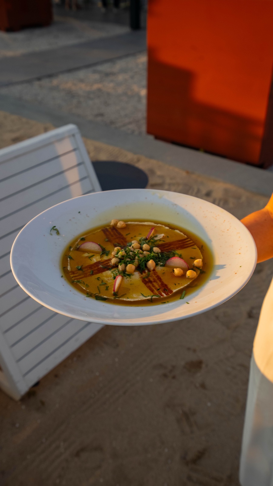 A plated BÁYRŪT dish garnished with chickpeas and herbs, carried to the table at sunset
